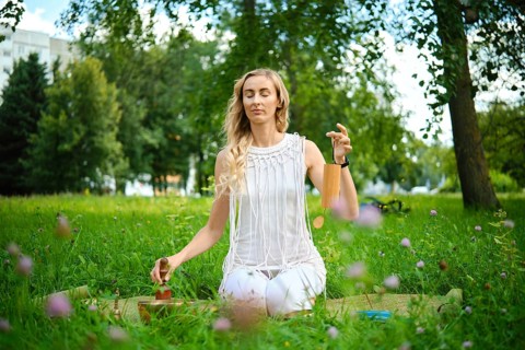 A person sits peacefully on a mat in a green park, gently playing singing bowls during meditation.