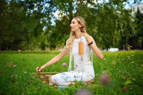 A woman sits calmly on a soft grassy area, softly striking a singing bowl while surrounded by