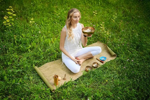 A woman sits cross-legged on a mat in a lush green field, holding a singing bowl. She is fully