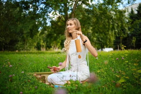 A woman sits cross-legged on the grass, surrounded by greenery, gently striking singing bowls with