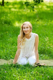 A woman with long hair sits on a yoga mat in a vibrant green park. She wears a light outfit and