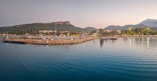 Kemer marina at dusk, filled with multiple docked boats. Lights from the harbor and hotels on the