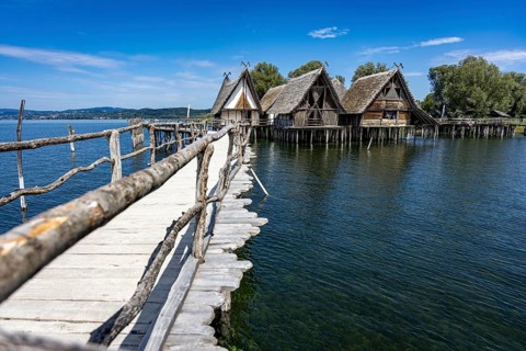 Wooden walkway leads to traditional stilt houses surrounded by calm water, pile dwellings
