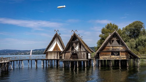 Three wooden pile dwellings with pointed roofs hover over calm waters, pile dwellings