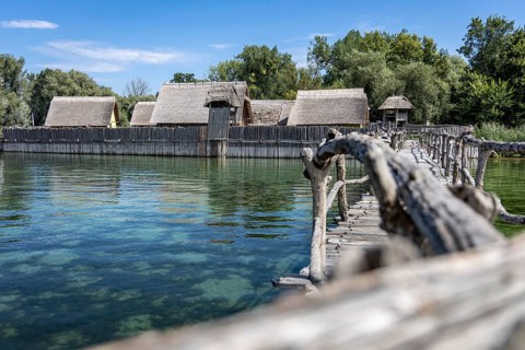 Wooden bridge stretches towards thatched buildings in the middle of idyllic nature, pile dwellings