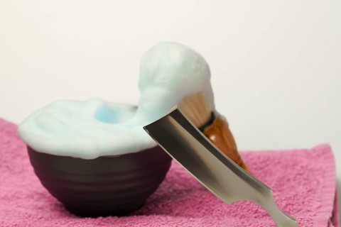 Shaving foam overflowing from a bowl with a straight razor and shaving brush resting on a pink