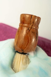 Wooden shaving brush dipped in light blue foam on a purple towel, evoking a sense of luxury and