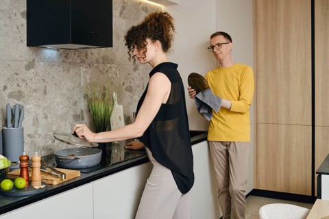 A woman stirs a pot on the stove while a a man wipes the washed dishes and watches her in a stylish