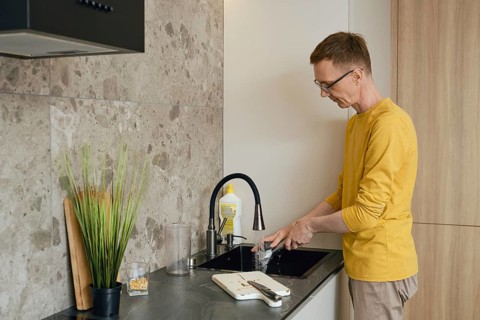 A person in a yellow long-sleeve shirt is washing dishes at a sleek kitchen sink. Sunlight