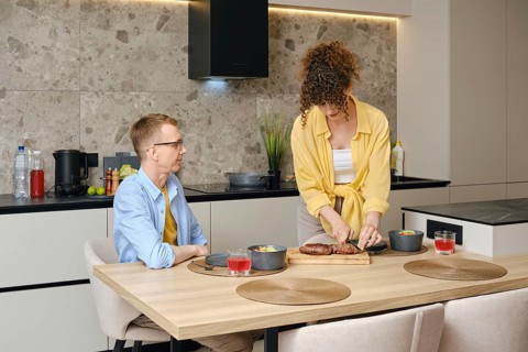 A woman slices roasted duck breast on a cutting board while a man sits at the dining table. They