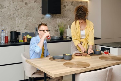 A man sits at a wooden dining table, sipping a drink, while a woman prepares a meal on the