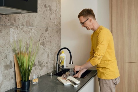 A man in a yellow long-sleeve shirt is washing utensils at a sleek kitchen sink. Natural light
