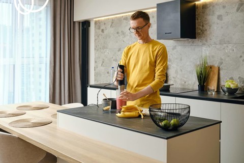 A middle-aged man prepares banana and strawberry smoothies in the kitchen using an immersion