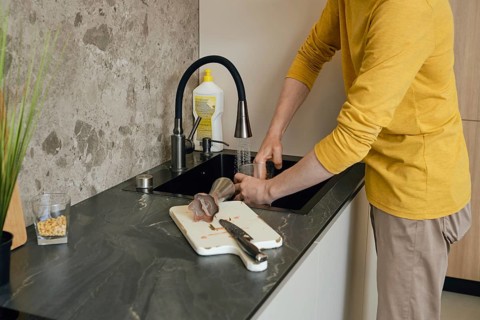 A person wearing a yellow shirt is washing utensils and a cutting board in a dark kitchen sink. The