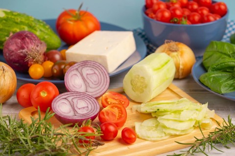 Fresh and sliced vegetables with feta on a wooden board, surrounded by basil and rosemary, rustic