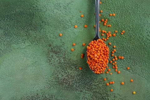 Bright orange lentils spill from a silver spoon onto a textured green surface, highlighting the