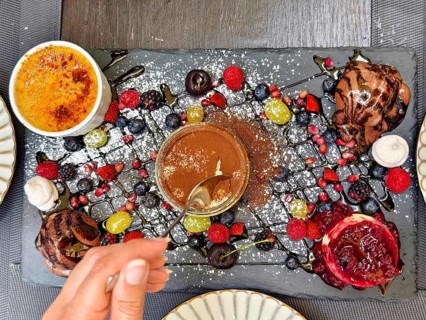 Hand with spoon tasting a dessert, surrounded by artfully arranged berries and chocolate Lower