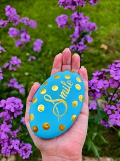 Hand holding blue stone with gold lettering and dots, purple flowers in the background Lower