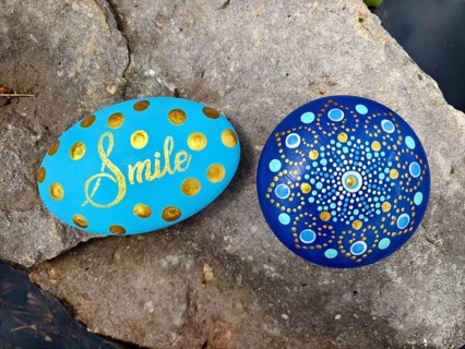 Two painted stones on rocks, one with 'Smile', one with a mandala pattern in blue and gold Lower