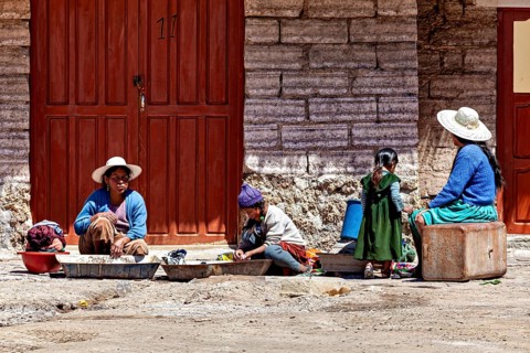 Women and children sit in front of a stone house, busy with housework. The sun shines brightly,
