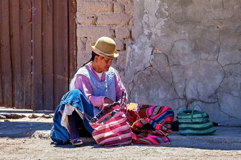 Man in traditional dress sits on the floor and sorts colourful bags on a wall, Traditional woman in