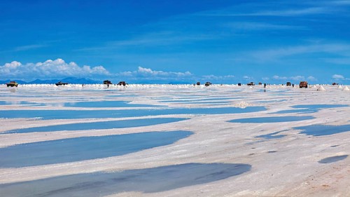 Puddles on a salt flat with vehicles on the horizon and clouds in the sky, The endless expanses of