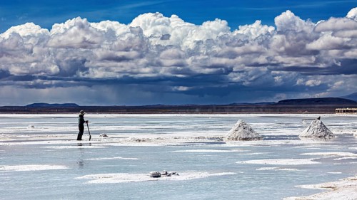 A single figure shovels salt under a dramatic cloudy sky on vast salt flats, The endless expanses