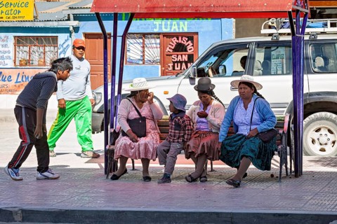 A group of people in traditional dress sit on a bench surrounded by shops and a car, people at a