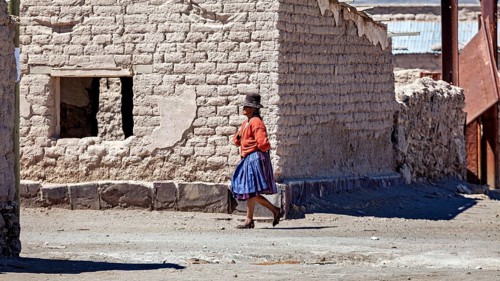 A woman in traditional dress walks along a dusty road in front of adobe houses, Traditional woman