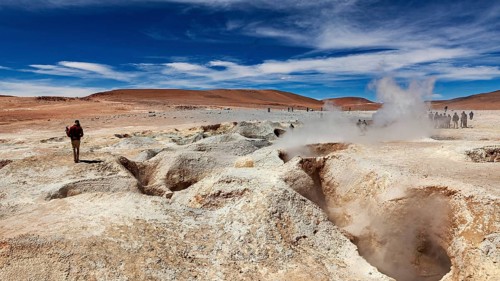 Lone hiker in a desert landscape with a smoking geyser and blue sky, The mud volcanoes and