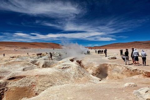 People standing in a desert landscape with a smoking geyser and blue sky, The mud volcanoes and
