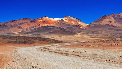 A winding sandy road through a dry landscape with reddish mountain peaks and blue skies, The
