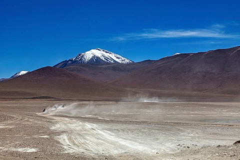 Vehicle tracks lead through a vast, dusty desert landscape to a snow-capped mountain, The landscape