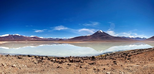 Wide view of a volcano with a reflecting lake and rocky shore under a blue sky, The Laguna Colorada