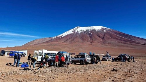 Groups of people and vehicles in a desert landscape with a snow-covered mountain under a blue sky,