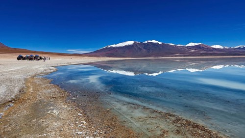 Panoramic view of a lake with snow-capped peaks, people and vehicles on the shore, The Laguna