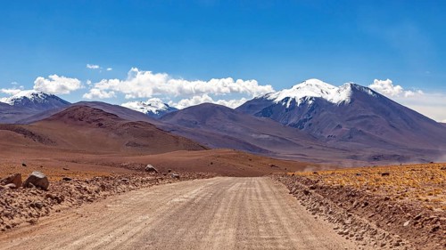 A straight, unpaved road leads through a barren mountain desert landscape on a sunny day, The