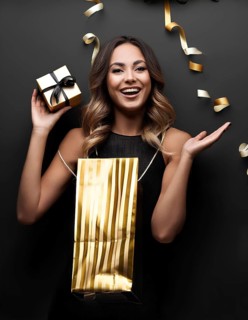 Smiling woman holding a small gift box and golden shopping bag with festive gold ribbons on a black