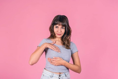 Young woman in gray t-shirt performing breast self-exam on pink background