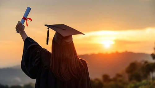 A graduate wearing a cap and gown raises a diploma with a red ribbon while standing outdoors at