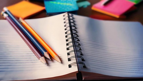 Three colorful pencils rest on an open spiral notebook with lined paper, surrounded by vibrant