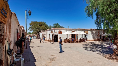 People sit under umbrellas on a lively street next to typical local buildings in the sunshine, The