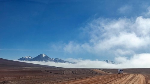 A dusty road leads through a remote desert landscape, bordered by mountains and clouds under a