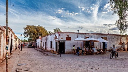 People move along a street with old buildings under partly cloudy skies and cyclists, The town of