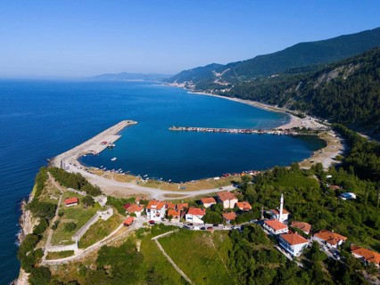 Aerial view of a coastal town with red roofs surrounded by green hills and calm blue sea, aerial