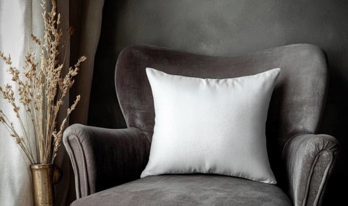 A white pillow sits on a gray chair in a room with a window. The pillow is placed on the back of