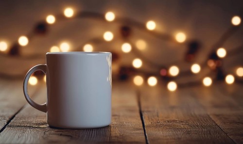 A white coffee cup sits on a wooden table with a string of lights in the background. The scene