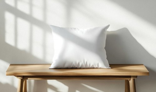 A white pillow sits on a wooden bench in front of a white wall. The pillow is empty and the bench