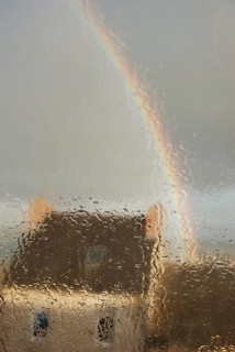 A house seen through a rain-soaked window with a rainbow arching across a gray sky, Old house