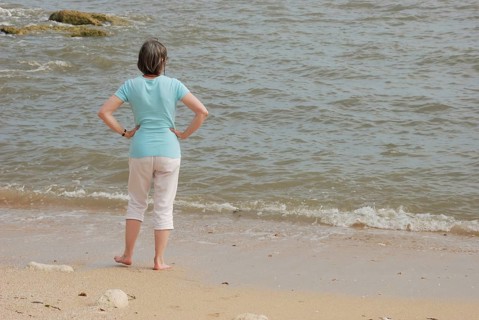 A woman stands on a sandy beach looking at the ocean waves, with rocks in the background in Royan